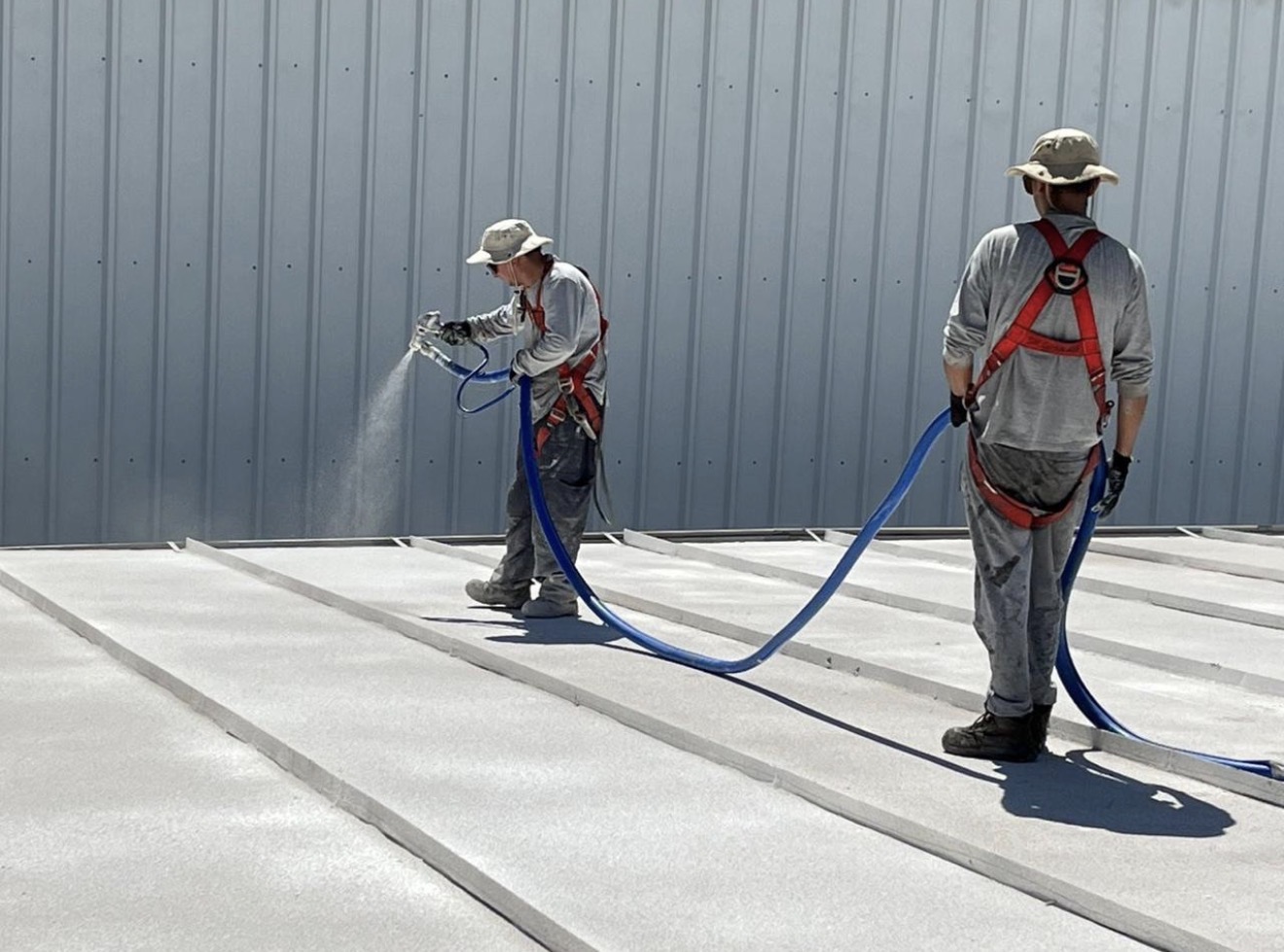 Two applicators spraying cork coating across a standing seam metal roof